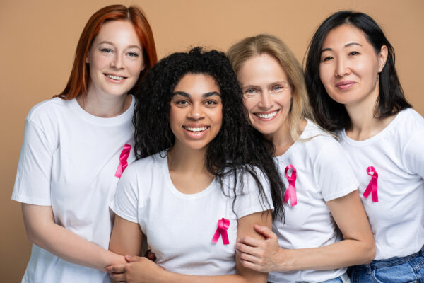 Portrait of smiling diverse group of multinational women with pink ribbon embracing looking at camera, isolated on clear background.